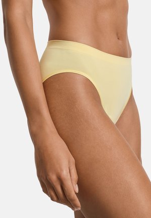 Vrouw die naadloos geel ondergoed met hoge taille draagt, afgebeeld van midden torso tot midden dij, met een ontspannen arm langs het lichaam tegen een witte achtergrond.