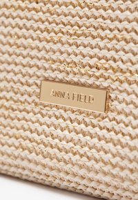 Zbliżenie na beżową, wypisaną fakturę z złotymi nićmi oraz prostokątną, złotą etykietą z wygrawerowanym napisem "ANNA FIELD".