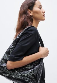 Čierna kabelka typu tote z lakovanej kože s textúrovaným vzorom, ktorú drží osoba oblečená v čiernom tričku, ukazujúca nadrozmerný dizajn a lesklý povrch.