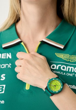 Grüne Uhr mit rundem Ziffernblatt, leuchtend gelbem Zifferblatt und schwarzen Akzenten. Wird an einer Hand getragen, die gegen ein tealfarbenes Poloshirt mit kontrastierenden Kragenstreifen gehalten wird.