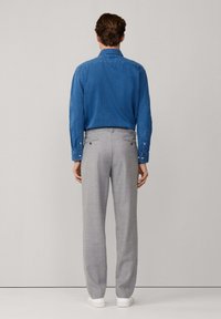 Uomo di spalle che indossa una camicia a maniche lunghe blu, pantaloni grigio chiaro e sneakers bianche su uno sfondo semplice.