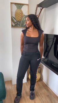 Žena s dlhými kučeravými vlasmi, oblečená v čiernom topu a čiernych nohaviciach, stojí vo vnútri pri televízii a rámovanej tlači ananásu.