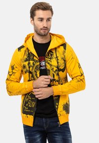 Sweat à capuche zippé jaune avec des motifs graphiques noirs, poches avant et cordon de serrage. En dessous, un t-shirt noir avec des lettres rouges.