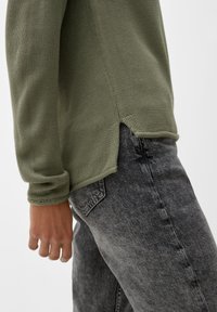 Pull en maille vert olive à manches longues et fentes latérales, associé à un jean denim gris clair avec une texture délavée et une poche arrière.