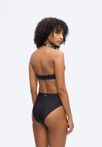 Γυναίκα με μπούκλες, ντυμένη με μαύρο μαγιό δύο τεμαχίων, γυρισμένη προς τα πίσω και κοιτάζοντας πάνω από τον ώμο της σε απλό φόντο.