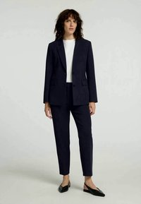 Kvinne med skulderlangt krøllete hår iført en marineblå dress, hvit topp og svarte spisse slingback-sko foran en ensfarget bakgrunn.
