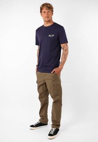 T-shirt en coton bleu marine avec un petit logo, associé à un pantalon cargo beige avec poches latérales, porté avec des baskets noires.