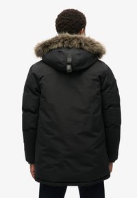 Czarna parka z futrzanym kapturem, wyposażona w tylną lotkę, zamek błyskawiczny i boczne kieszenie, zaprojektowana w celu zapewnienia ciepła i ochrony.
