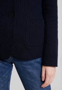 Marynarski żakiet w paski z teksturowanej tkaniny, z pojedynczym guzikiem i boczną kieszenią, zestawiony z niebieskimi dżinsami.