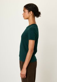 Temno zelena kratka majica s kratkimi rokavi, izdelana iz mehkega materiala, z okroglim izrezom in prilegajočim krojem. Vidni so žametni hlače s teksturo.