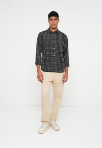 Man staat tegen een witte achtergrond en draagt een zwart-wit geruite lange mouwen overhemd, beige broek en off-white sneakers.