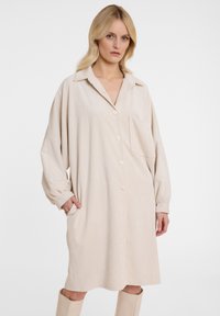Beiges Cord-Überhemdkleid mit langen Ärmeln, Knopfleiste vorne und seitlichen Taschen; verfügt über einen Kragen und eine einzelne Brusttasche.