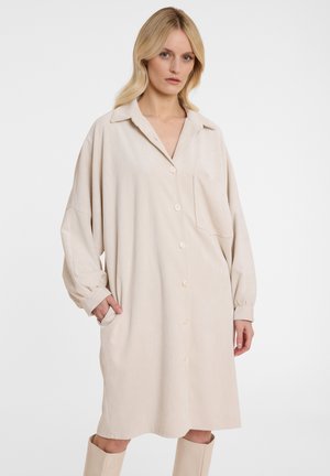 Beige ribfluwelen jurk met lange mouwen, knoopsluiting aan de voorkant en zijzakken; heeft een kraag en een enkele borstzak.