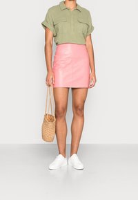 Chemise à manches courtes vert olive, mini-jupe en cuir rose, baskets blanches, tenant un sac panier tissé. Lignes épurées et textures lisses.