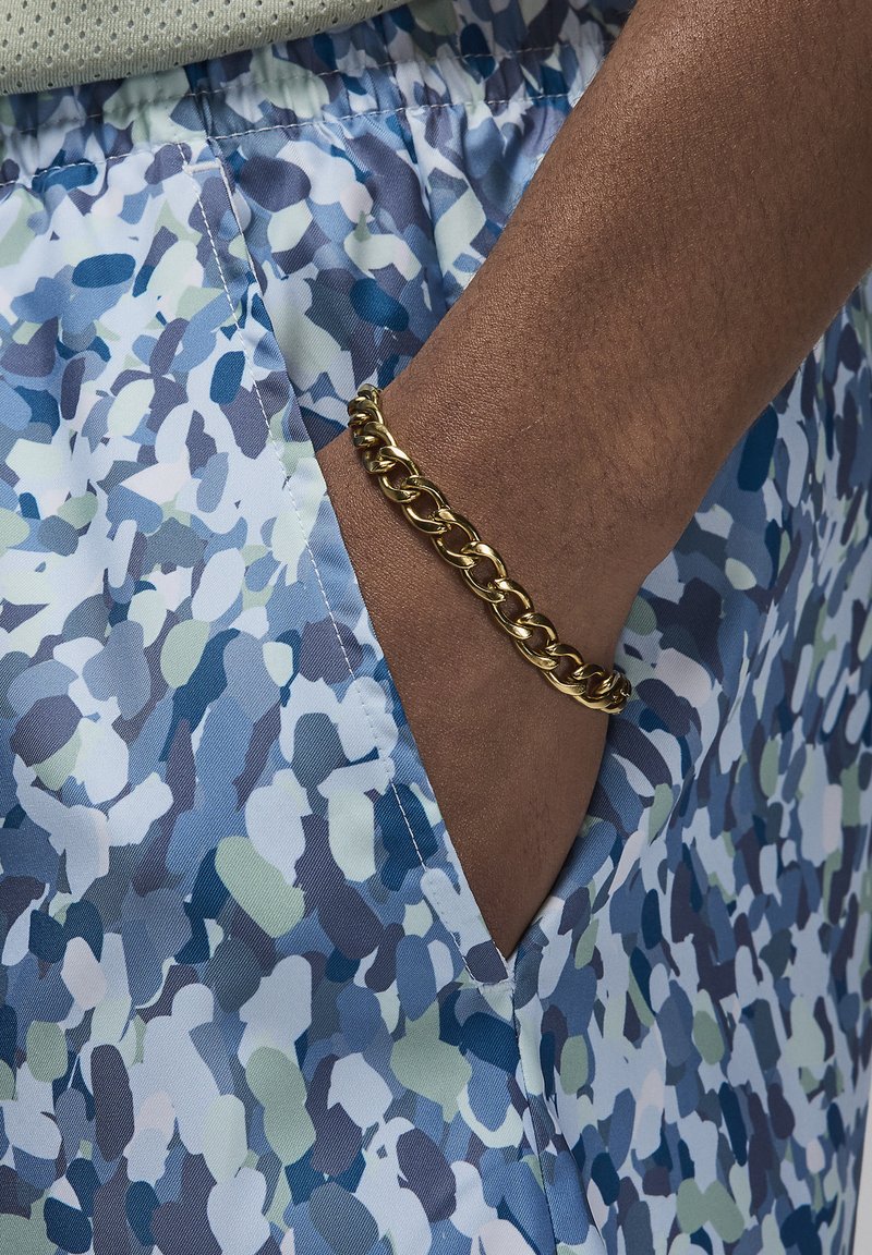 Shorts multicolores présentant un motif abstrait bleu, vert et blanc, avec une taille élastique et une poche visible. Un bracelet en chaîne doré orne le poignet.