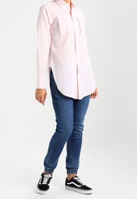 Camicia a maniche lunghe di colore rosa chiaro con colletto e due tasche sul petto, abbinata a jeans blu a gamba affusolata e sneaker nere con dettagli bianchi.