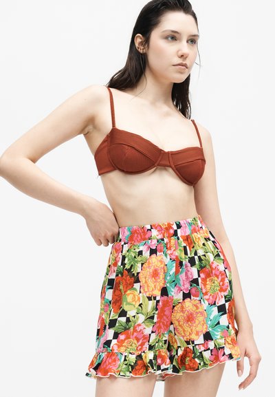 Νεαρή γυναίκα φοράει τοπ γραμμωτό σε σκουριά χρώμα και σόρτς με λουλούδια και βολάν στο τελείωμα, στέκεται με το χέρι στη μέση.