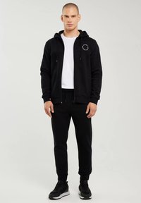 Bărbat în picioare purtând hanorac negru cu fermoar, tricou alb, pantaloni de trening negri și adidași negri, pe un fundal simplu și deschis la culoare.