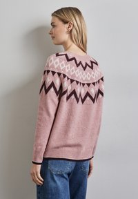 Rosa stickad tröja med ett geometriskt mönster i mörkbrunt och vitt, med ribbade ärmslut och nederkant, visad från sidan mot en enkel bakgrund.