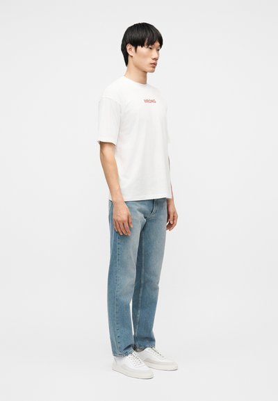 Jeune homme debout portant un T-shirt blanc avec le texte "WRONG", un jean bleu clair et des baskets blanches, sur un fond blanc uni.