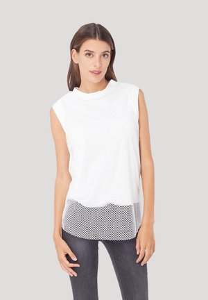 Top sin mangas de malla blanca con cuello redondo y dobladillo con volantes, superpuesto sobre una base de tela sólida. Combinado con jeans gris oscuro.