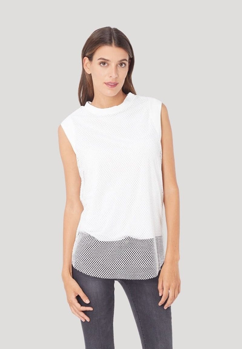 Top sin mangas de malla blanca con cuello redondo y dobladillo con volantes, superpuesto sobre una base de tela sólida. Combinado con jeans gris oscuro.