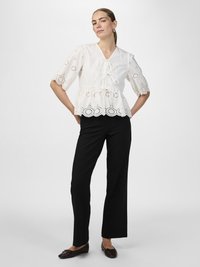 Blusa peplum bianca con dettagli ad occhio di pernice, scollatura a V e fiocco. Abbinata a pantaloni neri a gamba dritta e scarpe basse scure.