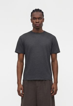 Νέος άνδρας με πλεξούδες στα μαλλιά, φορούν απλό σκούρο γκρι κοντομάνικο μπλουζάκι και φαρδιά σκούρα καφέ παντελόνια σε λευκό φόντο.