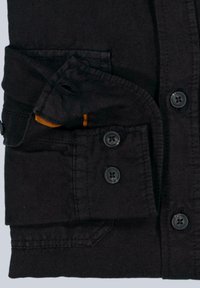 Schwarzes Hemd mit Knopfleiste, zwei Brusttaschen und hochgekrempelten Ärmeln. Weicher Stoff und kontrastierendes oranges Detail in einer Tasche.