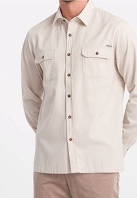 Camisa de manga larga color beige claro con cierres de botones marrones, dos bolsillos en el pecho y un corte relajado. Tejido de mezcla de algodón con una textura suave.