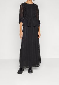 Abito maxi in pizzo nero con maniche lunghe, dettagli arricciati e un design testurizzato. Abbinato a robusti scarponi neri.