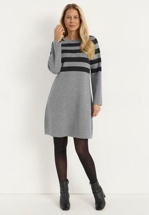 Frau in einem knielangen grauen Kleid mit schwarzen horizontalen Streifen auf der oberen Brust und den Ärmeln, trägt schwarze Strumpfhosen und schwarze Stiefeletten.