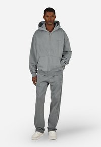 Camisola cinza com fecho de correr e capuz, bolsos frontais e calças cinzentas a condizer. O tecido parece macio com um ajuste confortável; os sapatos são brancos.