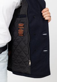 Granatowy płaszcz wełniany z podszewką z czarnego pikowanego materiału. Posiada zamek błyskawiczny, guziki oraz pomarańczową metkę z napisem "BOMBERS". Widoczne kontrastowe szwy.