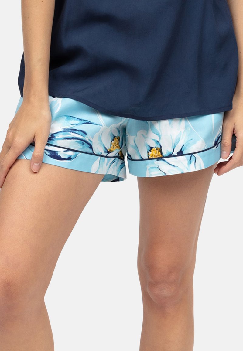 Vrouw in lichtblauwe bloemen shorts en een marineblauwe top, met blote benen en handen tegen een effen achtergrond.