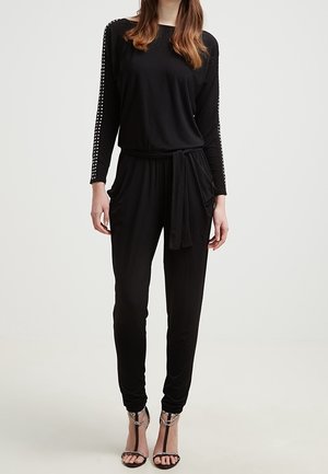 Femme portant une combinaison noire à manches longues avec des manches cloutées, une ceinture nouée à la taille et des sandales à talons noirs ornées de détails en chaîne.