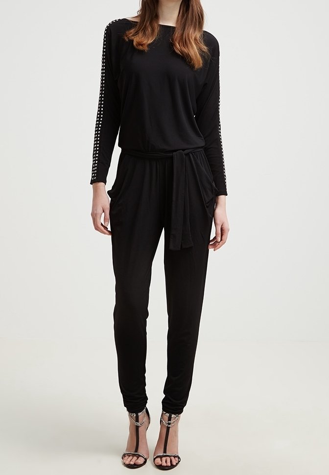 Femme portant une combinaison noire à manches longues avec des manches cloutées, une ceinture nouée à la taille et des sandales à talons noirs ornées de détails en chaîne.