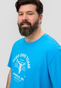 Męska turkusowa koszulka z bawełny, z białym nadrukiem graficznym, który zawiera napis "CLIFF DIVE TEAM", z postacią nurka i falami.