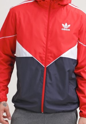 Rode, witte en marineblauwe Adidas jas met rits en logo op de borst, gedragen door een persoon met een getatoeëerde hand en grijze broek.