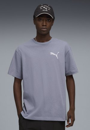 Jong persoon die een lichtgrijs Puma T-shirt met een wit logo draagt en een zwarte Puma Athletics pet op heeft, staand tegen een grijs verloopachtergrond.