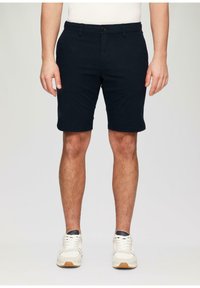 Pantalones cortos de color náutico fabricados en algodón, con una parte delantera plana, trabillas para cinturón y un cierre de botón único. Combinados con zapatillas blancas.