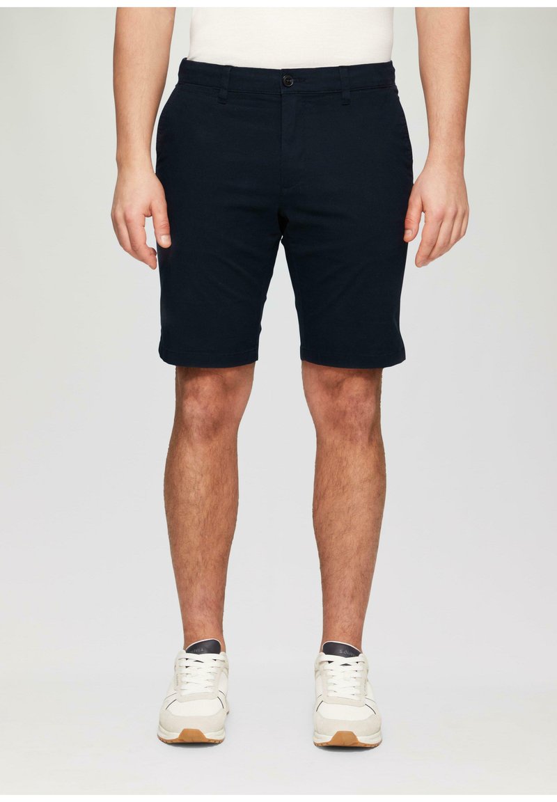 Pantalones cortos de color náutico fabricados en algodón, con una parte delantera plana, trabillas para cinturón y un cierre de botón único. Combinados con zapatillas blancas.