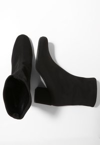 Par de botas de ante negras hasta la mitad de la pantorrilla con tacón cuadrado, una vista desde arriba y la otra de lado sobre un fondo blanco.