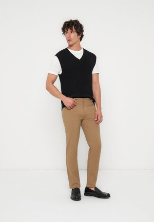 Jeune homme aux cheveux bouclés portant un pull noir sans manches par-dessus un t-shirt blanc, un pantalon beige et des mocassins noirs, debout contre un mur blanc.