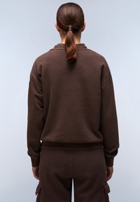 Een bruine sweatshirt met een relaxte pasvorm, geribbelde kraag en manchetten, zachte textuur en een eenvoudig ontwerp. Gemaakt met bijpassende bruine broek.