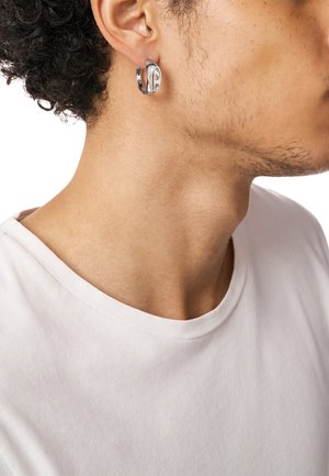 Hopeinen rengaskorvakoru, jossa on tyylikäs muotoilu, sileät pinnat ja pieni pyöreä leikkaus. Päällä vasemmassa korvassa, yhdistetty valkoiseen paitaan.