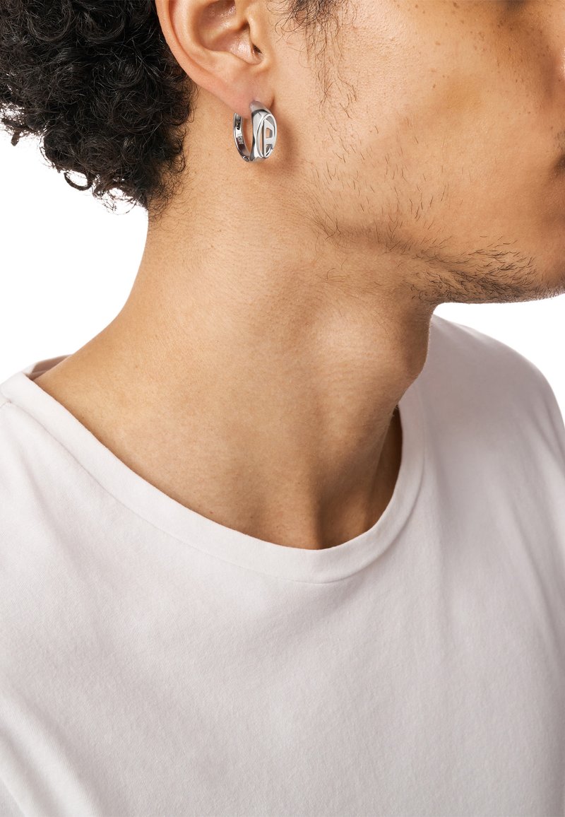Boucle d'oreille argentée en forme de créole au design épuré, présentant des surfaces lisses et une petite découpe circulaire. Portée à l'oreille gauche, assortie à une chemise blanche.