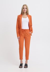 Blazer naranja y joggers a juego hechos de una tela suave, que cuentan con una cintura ajustable con cordón. Llevado con una camiseta sin mangas blanca y sandalias negras tipo slide.