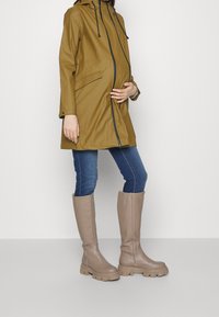 Impermeabile khaki con cappuccio, zip frontale e tasche; abbinato a stivali beige fino al ginocchio con suola spessa e parte superiore testurizzata.