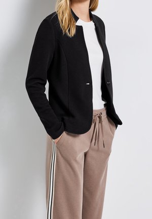 Frau trägt schwarzen strukturierten offenen Blazer, weißes T-Shirt und beige Kordelzughose mit schwarz-weißen Seitenstreifen, Hände in den Taschen.